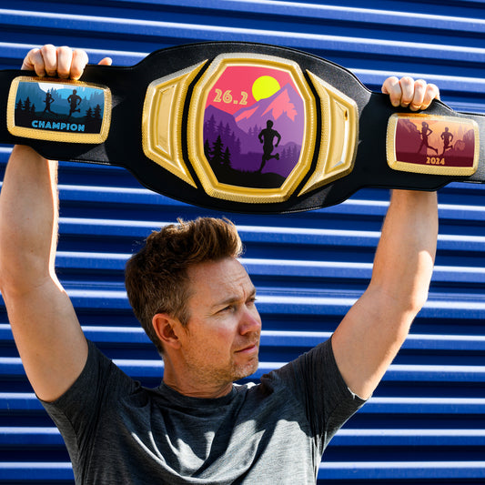 A man holds a WWE-inspired championship belt over his head. The design on the belt has been customized by TrophySmack to celebrate completing a 26.2 mile running race.